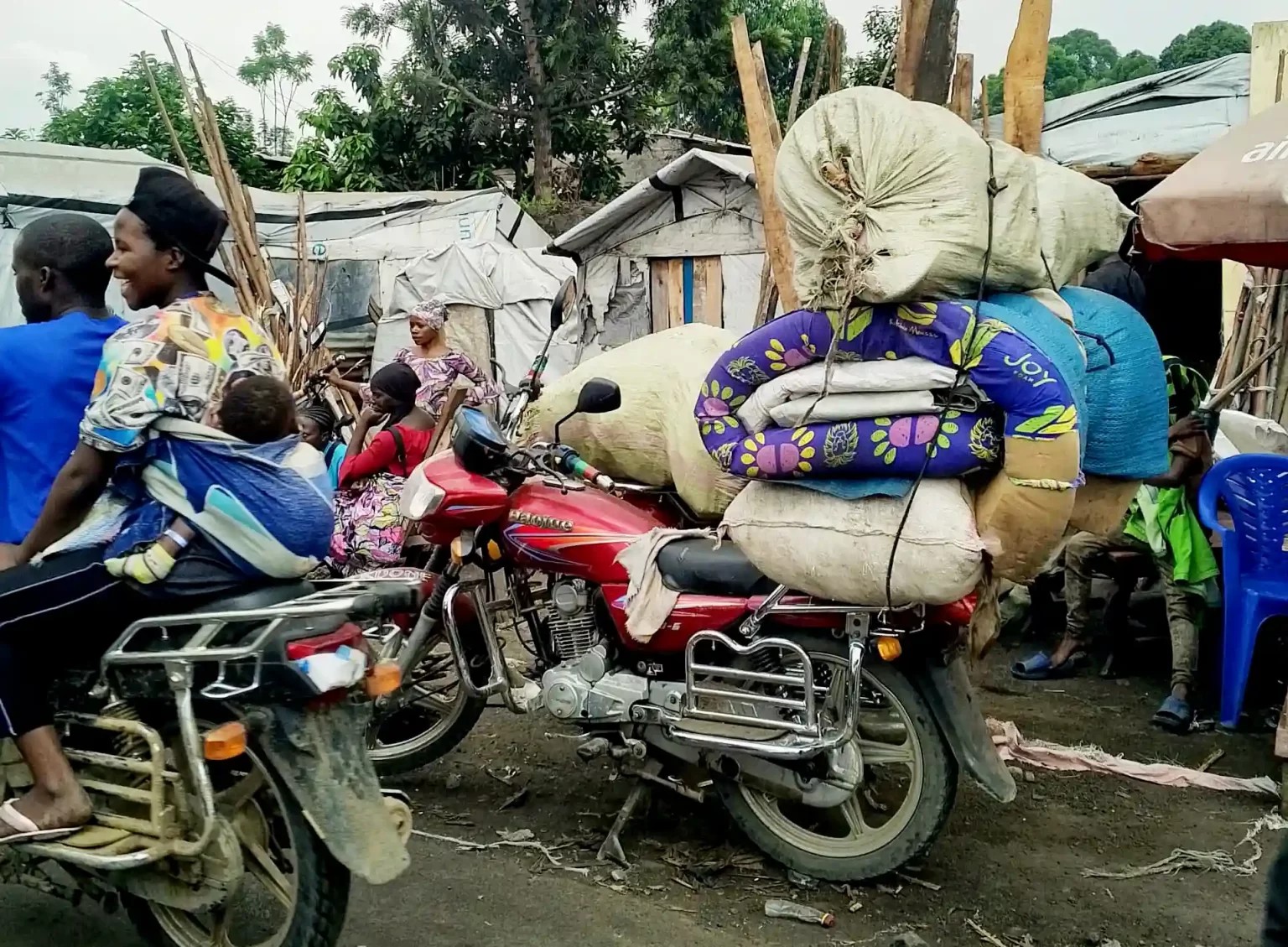 Ein Motorrad transportiert Matratzen, auf einem anderen sitzt eine Frau mit Baby. Im Hintergrund Häuser.